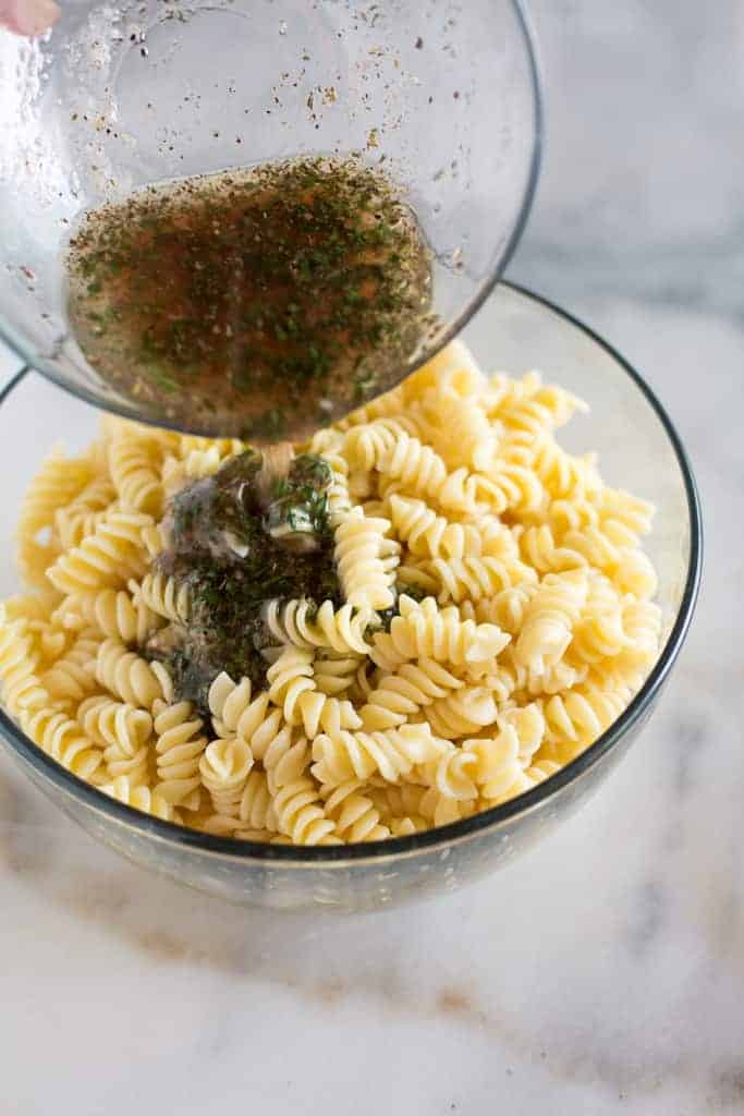 Homemade Italian pasta salad dressing being poured over a bowl full of cooked rotini pasta.