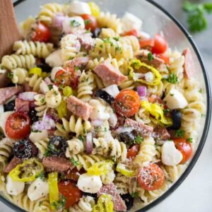Cold Italian pasta salad with rotini noodles, mozzarella, tomatoes, olives, and salami in a clear glass bowl.