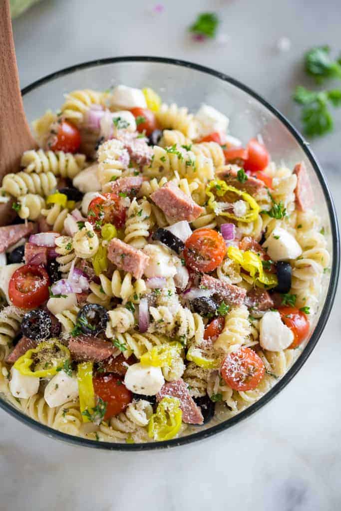 Cold Italian pasta salad with rotini noodles, mozzarella, tomatoes, olives, and salami in a clear glass bowl.