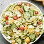 A bowl full of pesto pasta salad with farfalle noodles, red and yellow cherry tomatoes, mozzarella, cucumbers, tossed in pesto sauce.