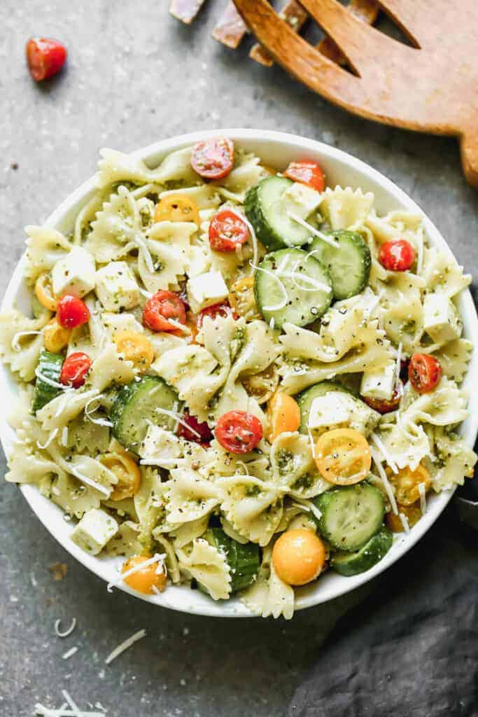 A bowl full of pesto pasta salad with farfalle noodles, red and yellow cherry tomatoes, mozzarella, cucumbers, tossed in pesto sauce.