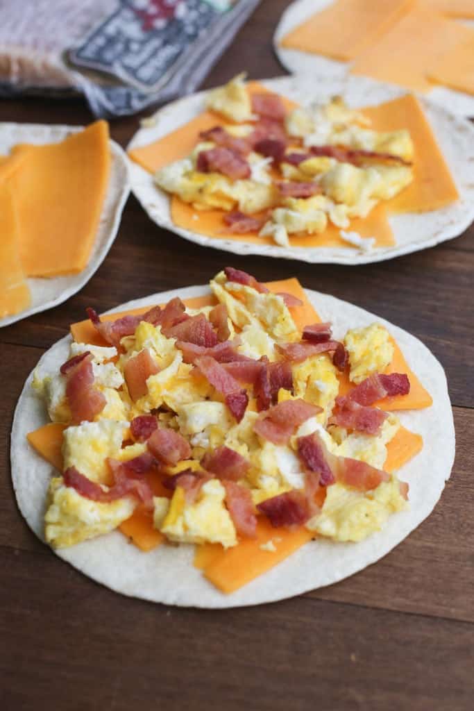 A tortilla topped with cheddar cheese slices, scrambled eggs, and bacon.