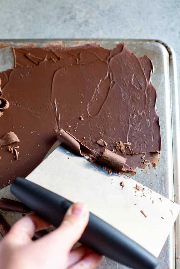 A hand holding a metal scraper to scrape chocolate up from a baking sheet to form chocolate curls.