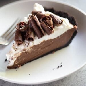 A slice of chocolate cream pie with whipped cream and chocolate curls on top, on a white plate with a fork.