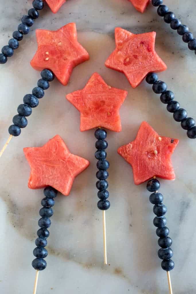 Fruit kebabs on a white marble board, called fruit sparklers, with blueberries and a watermelon star secured on a wood skewer.