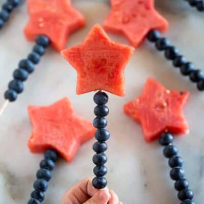 Two hands holding a wooden skewer lined with blueberries and a watermelon cut into the shape of a star, on the top.