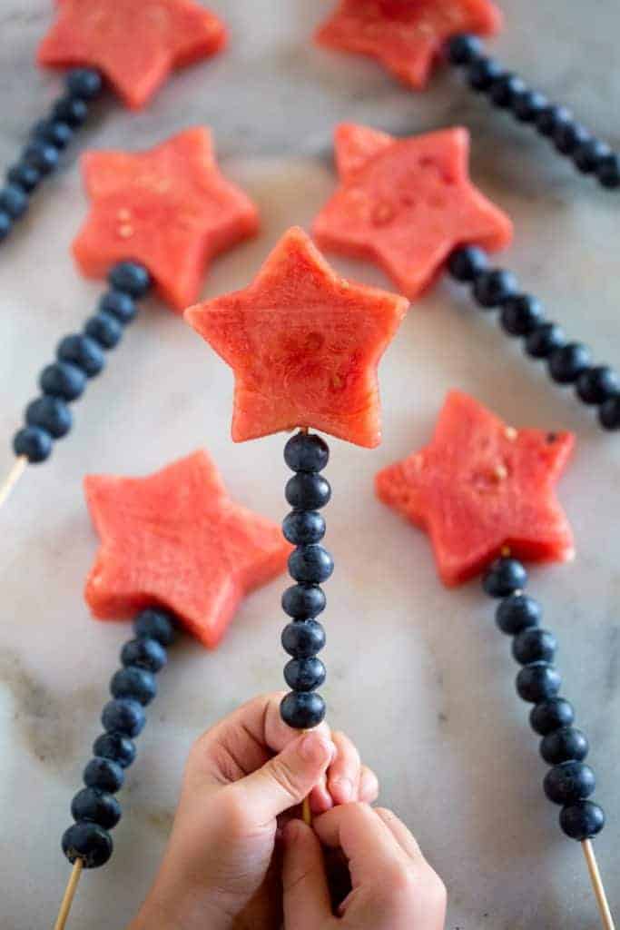 Two hands holding a wooden skewer lined with blueberries and a watermelon cut into the shape of a star, on the top.