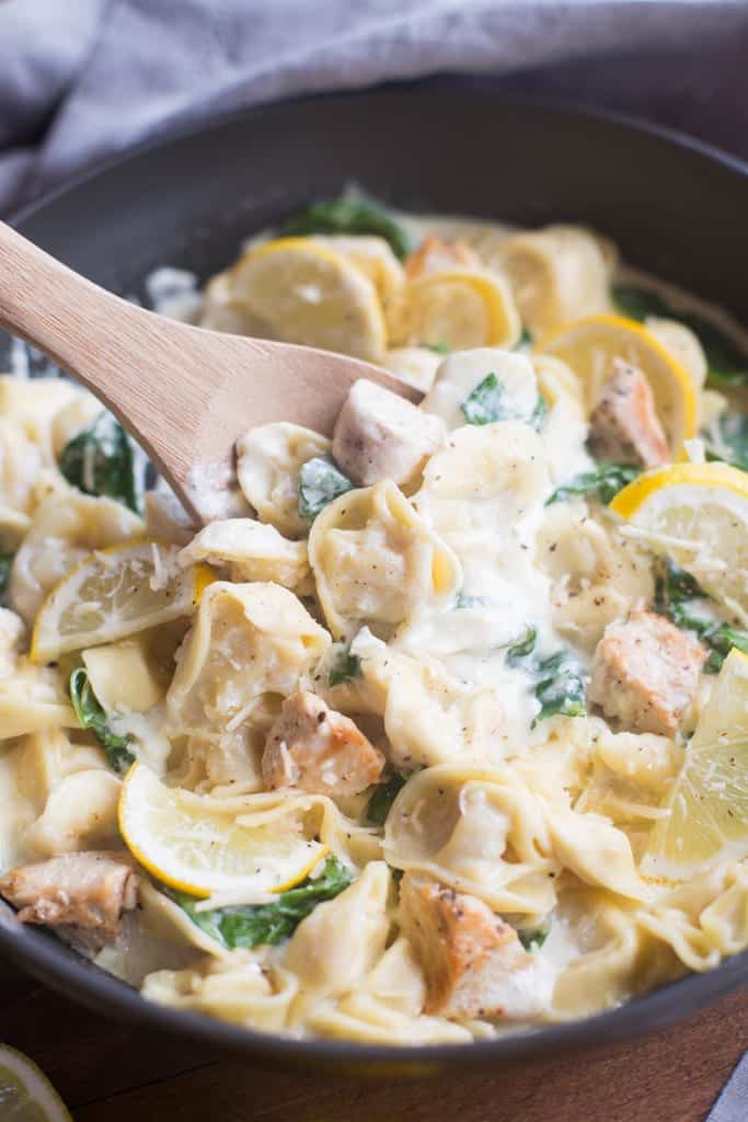 A wooden spoon stirring tortellini, chicken, spinach, and lemon slices in a creamy sauce.