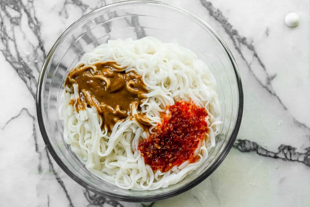 Cooked rice noodles in a glass mixing bowl with peanut sauce and sweet chili sauce on top.