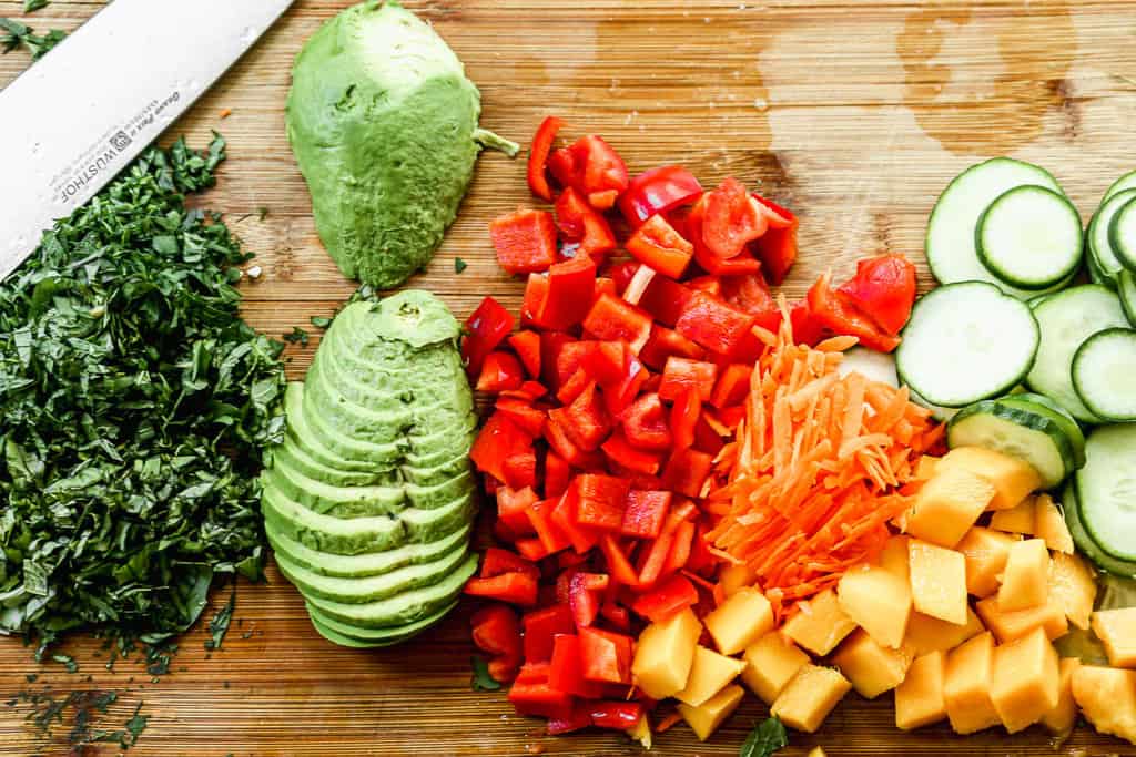 Sliced avocado, bell pepper, carrots, cucumber, mango, and fresh herbs all sliced on a wooden cutting board.