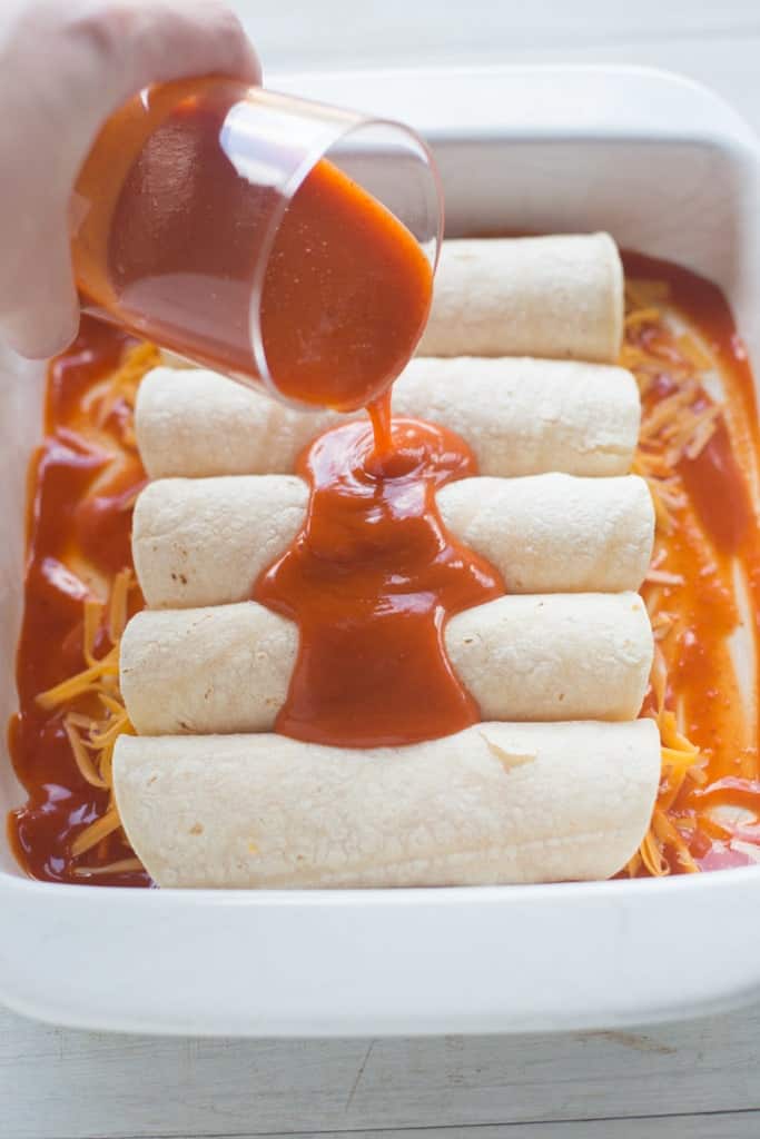 A white casserole dish filled with five uncooked cheese enchiladas with red enchilada sauce being poured over the top.
