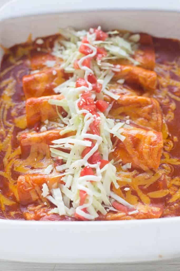 A casserole dish filled with a row of cheese enchiladas and topped with red enchilada sauce, shredded cabbage, tomatoes, and cheddar cheese.