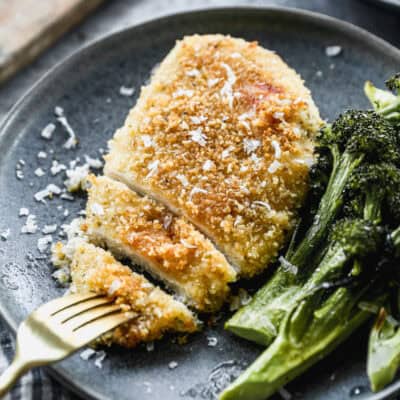 A piece of baked Parmesan Crusted Chicken on a plate served with broccolini, with a fork picking up a slice to eat.