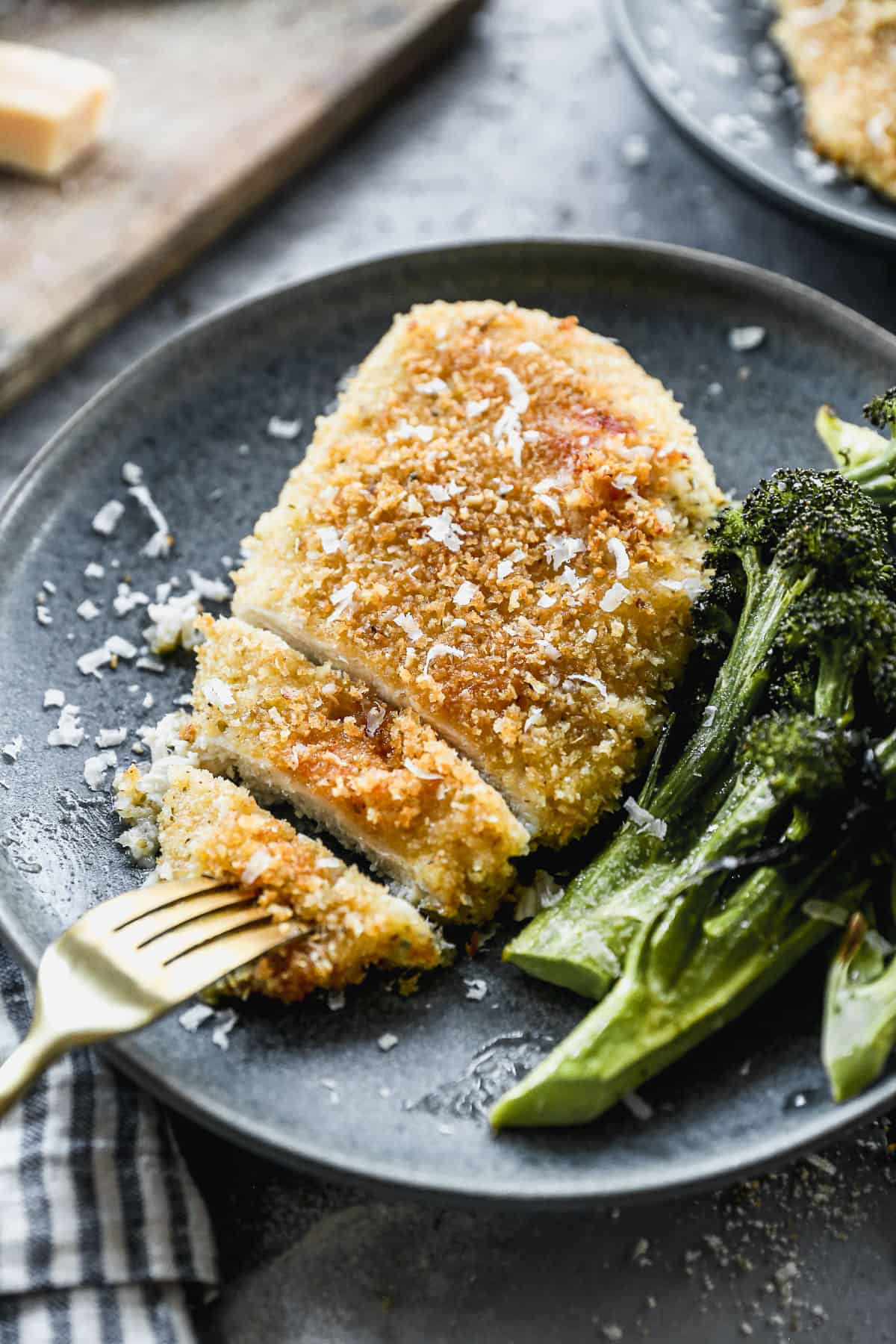 A piece of baked Parmesan Crusted Chicken on a plate served with broccolini, with a fork picking up a slice to eat.