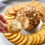 Cheese ball being spread on a cracker with a plate of crackers and the rest of the cheeseball in the background.