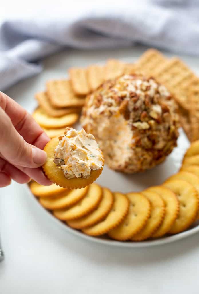 Cheese ball being spread on a cracker with a plate of crackers and the rest of the cheeseball in the background.