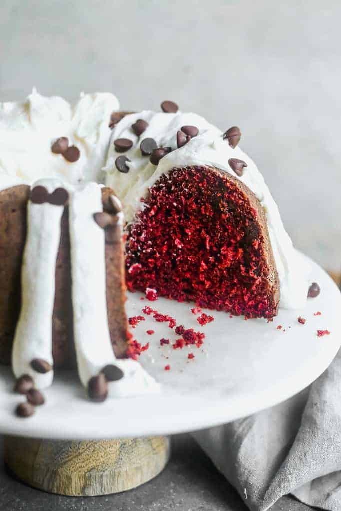 A Red Velvet Bundt Cake with cream cheese frosting on a cake stand, with a piece cut from it.