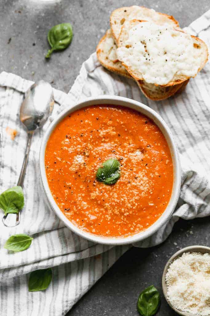 Roasted tomato soup served in a white bowl with slices of cheesy bread on the side.
