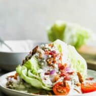 A wedge salad on a plate, topped with blue cheese dressing, bacon crumbles, tomato, and onion.