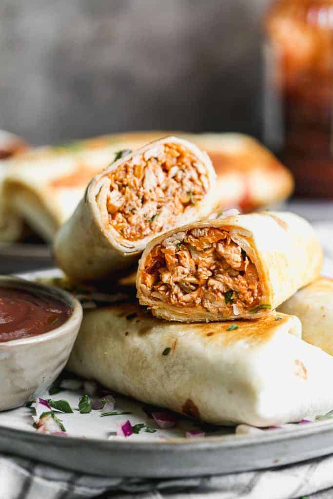 A BBQ chicken wrap cut in half to show the filling inside the tortilla, stacked with other wraps on a plate. 