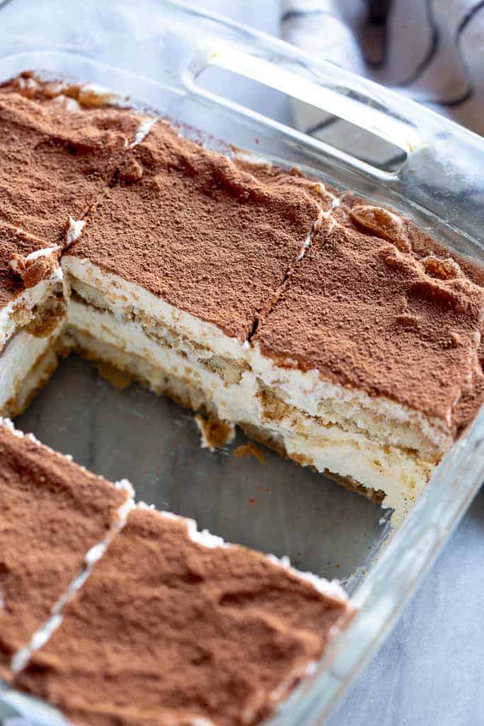 A clear glass pan of tiramisu with two slices removed from the center.