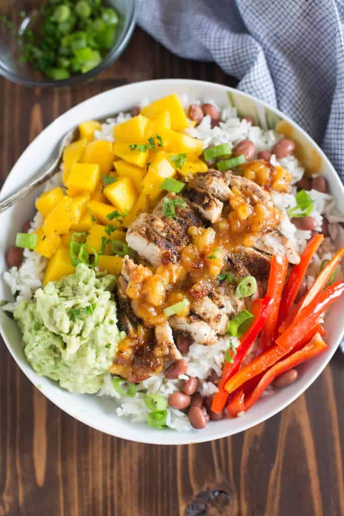 A bowl filled with jerk seasoned chicken, mangos, guacamole, bell peppers, and beans over rice. 