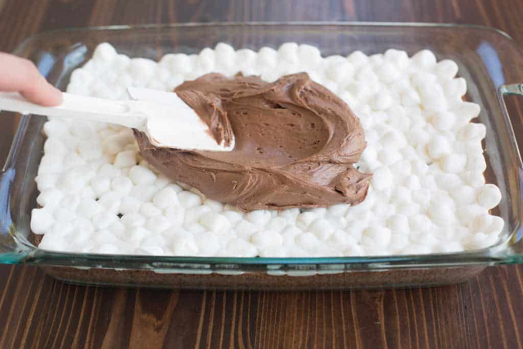 Chocolate frosting being spread on top of miniature marshmallows, which cover a pan of brownies.