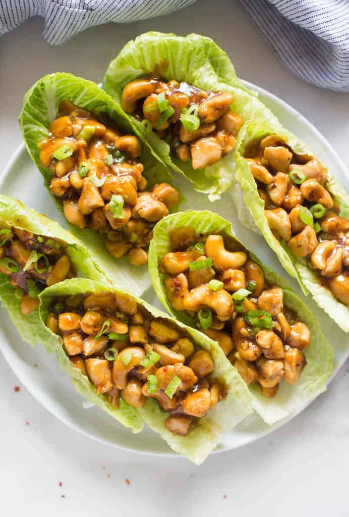 Six lettuce leaves filled with cashews, chicken, and a homemade sauce, all on a white plate. 