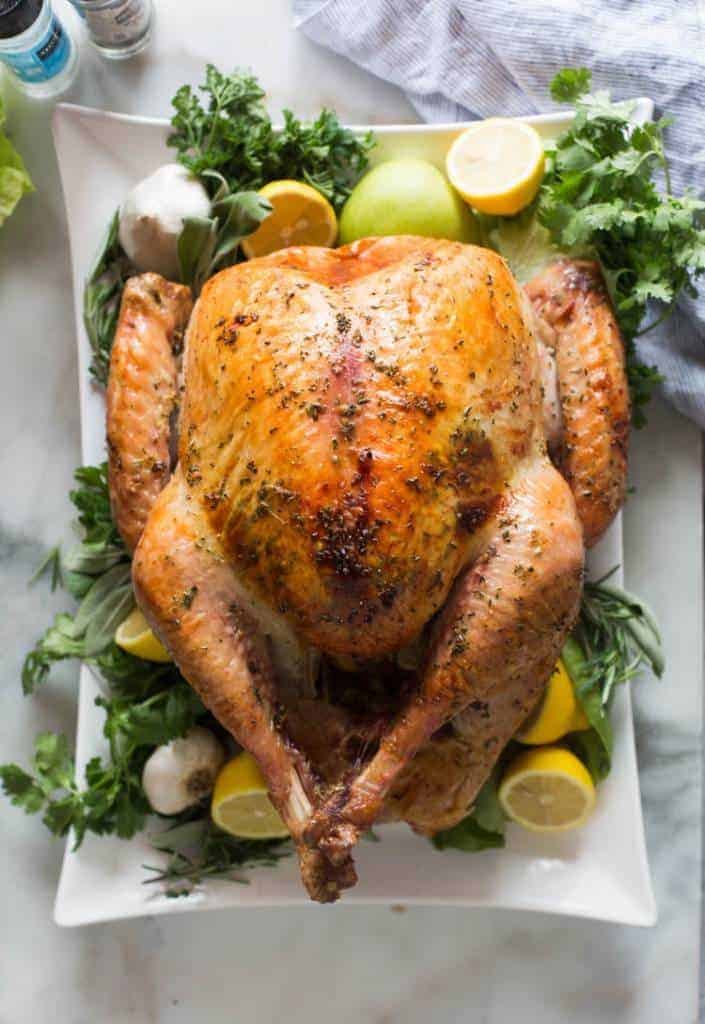 Roasted Turkey on a large white platter, garnished with lemons, apples, garlic and fresh green herbs.
