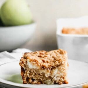 A slice of Apple Coffee Cake on a white plate with the cake pan and a bowl of apples in the backround.