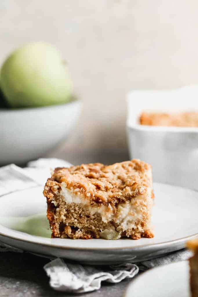 A slice of Apple Coffee Cake on a white plate with the cake pan and a bowl of apples in the backround.