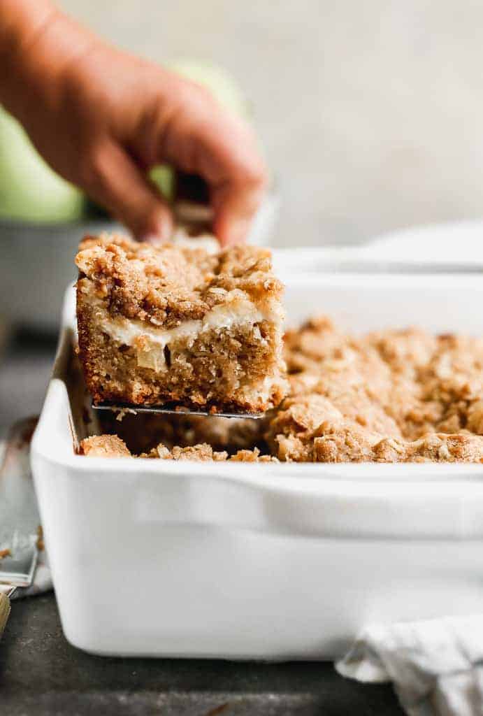 A pan of baked Apple Coffee Cake with a hand and spatula lifting a piece from the pan.