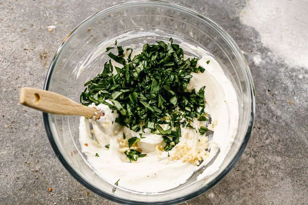 A bowl with cream cheese, ricotta, sour cream, basil and garlic in a mixing bowl.