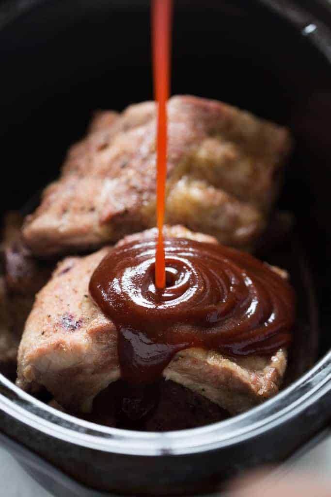 A rack of pork baby back ribs in a slow cooker with barbecue sauce being poured on top.