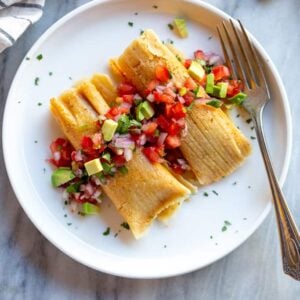 Two tamales on a plate with pico de gallo and chopped avocado.