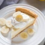 Banana Cream Pie slice on a white plate with fresh banana slices on top of whipped cream.