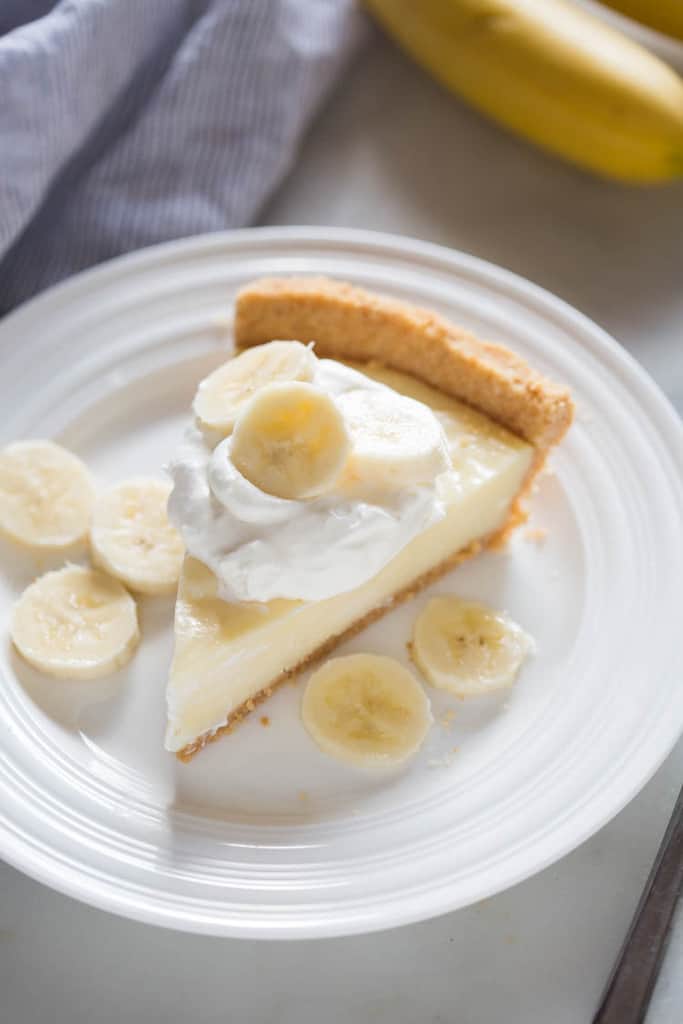Banana Cream Pie slice on a white plate with fresh banana slices on top of whipped cream.
