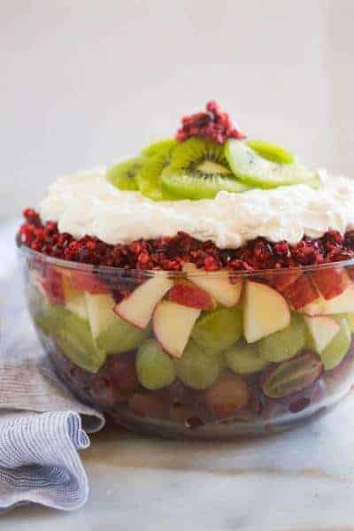 Side profile of a clear bowl filled with fruit including red and green grapes, apples, cranberries, whipped cream and kiwi.