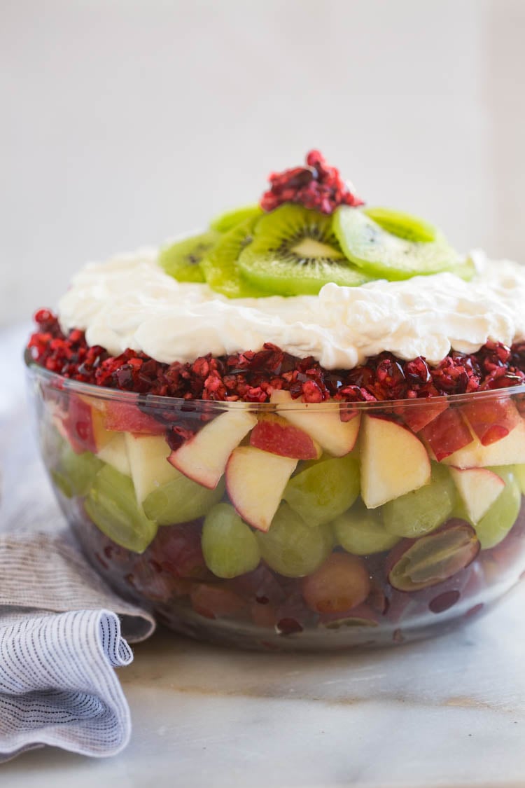 Side profile of a cranberry apple fruit salad layered in a clear bowl with red and green grapes, whipped cream with vanilla yogurt, and kiwi.