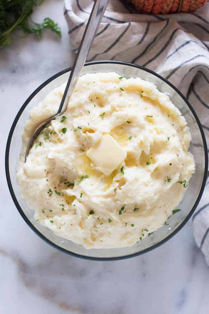 An overhead view of a clear bowl filled with mashed potatoes, melted butter, parsley and a silver serving spoon.