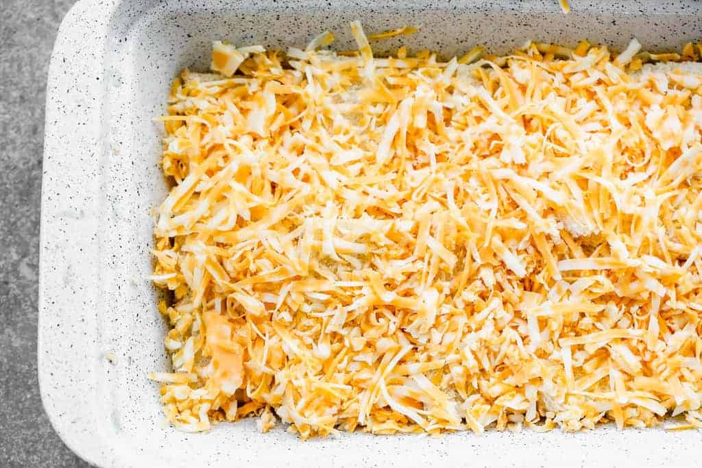 Shredded cheese spread over bread slices in a casserole dish.