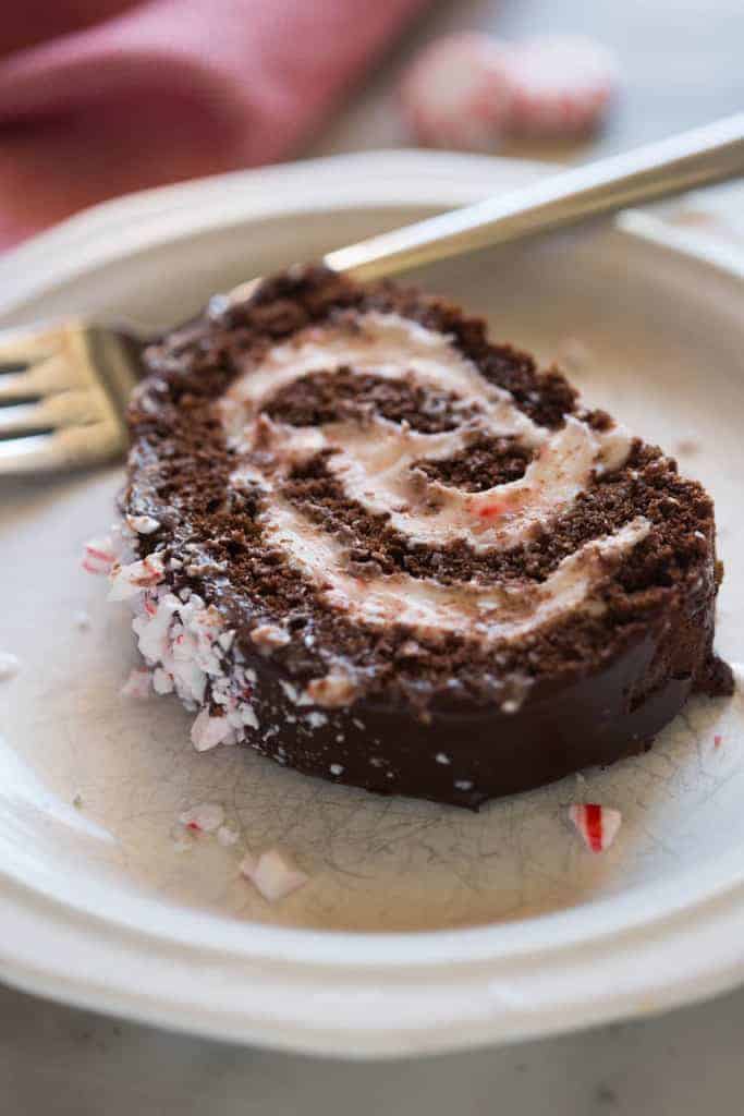 A piece of chocolate peppermint cake roll on a white plate with a fork and red towel and peppermint candies in the background.