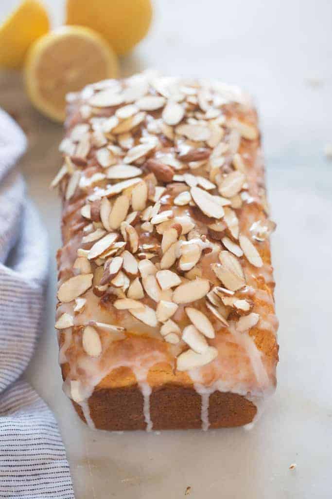 A loaf of lemon almond bread with glaze and sliced almonds covering the top of the loaf, resting on a marble board with a blue towel next to it and lemons in the top corner of the photo.