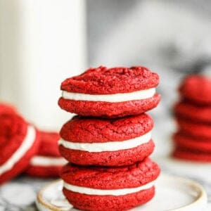 Three red velvet cookies with cream cheese frosting, stacked on top of each other on a plate.