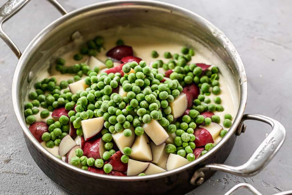 Cooked red potato bites and green peas added to a white cream sauce in a skillet.