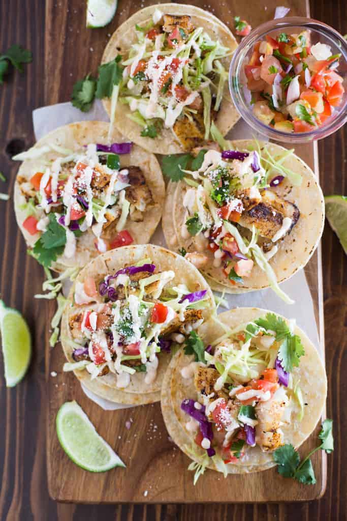 Overhead photo of fish tacos served on a wood board, on corn tortillas with toppings including cabbage and pico de gallo.