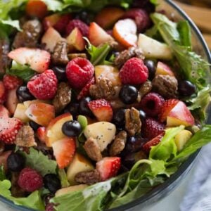 Mixed green salad with strawberries, blueberries, raspberries, apple, onion, feta cheese and poppyseed dressing in a glass bowl with salad tongs in the background.