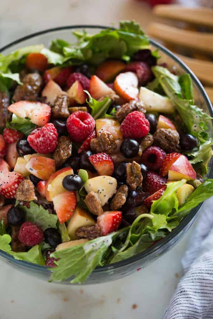 Mixed green salad with strawberries, blueberries, raspberries, apple, onion, feta cheese and poppyseed dressing in a glass bowl with salad tongs in the background. 