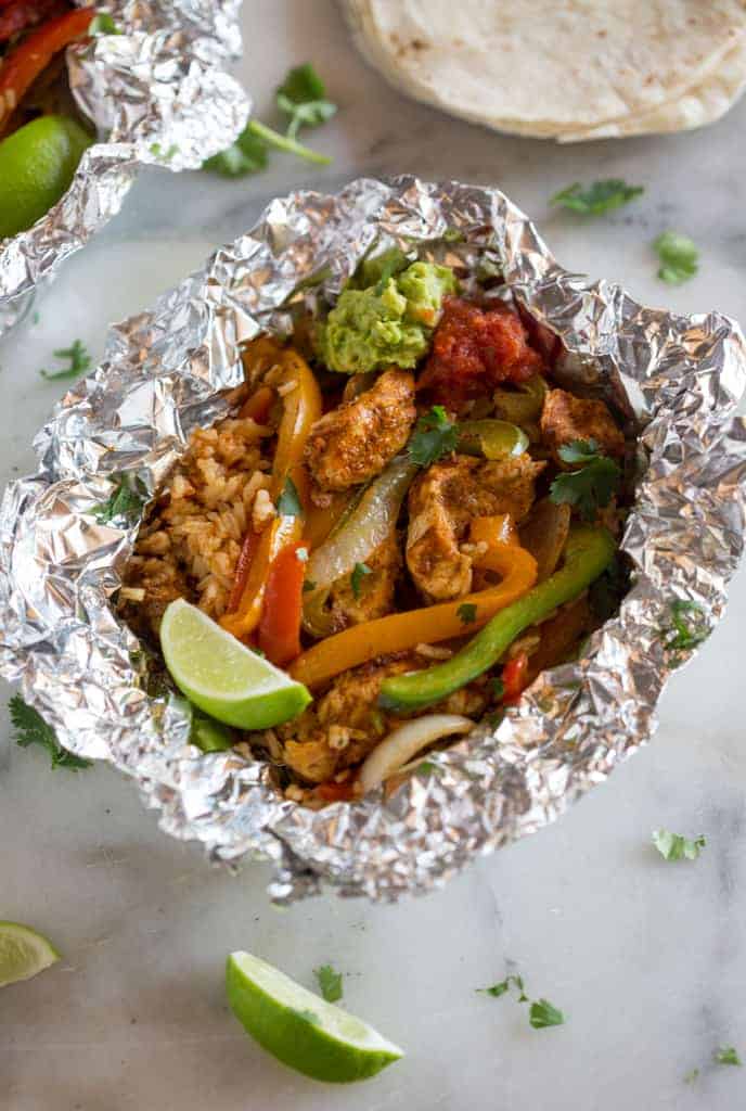 An opened foil packet dinner with chicken, rice, bell peppers, onion, lime and cilantro, with flour tortillas in the background.