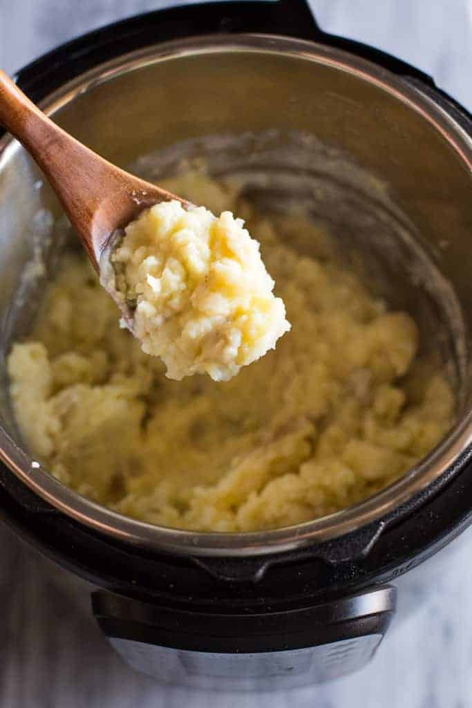 Mashed potatoes inside an instant pot with a wooden spoon lifting a spoonful of potatoes from the pot.